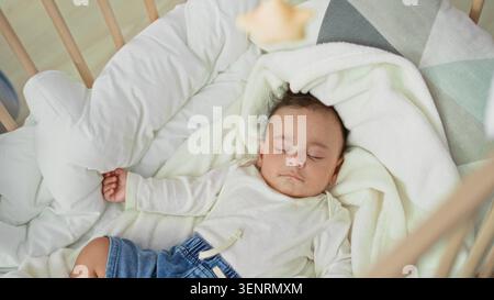 Hispanischer Kleinkind, der auf dem Rücken in einem hölzernen Wiegenbett mit weißer Decke und Jeansshorts schläft, Hand auf Kissen im Kinderzimmer gerollt; friedliche Ruhe. Stockfoto