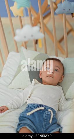 Hispanischer Junge, der im Kinderbett auf dem Bett liegt, Kopf auf gemustertem Kissen, trägt Jeansshorts und blickt auf das Plüschmobil, das über Holzlatten hängt Stockfoto