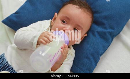 Baby Boy hält Milchflasche und stillt, während er auf dem Bett im Gebäude weint; verstörte beruhigende Komfortpflege. Stockfoto