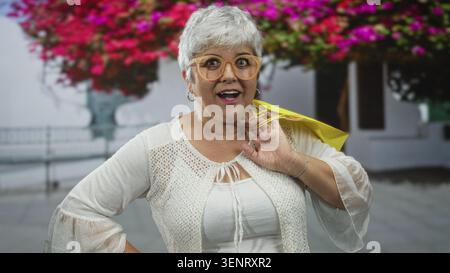 Seniorin mit gelber Einkaufstasche und Hand auf der Straße mit Gesten, Brille und Strickjacke neben Blumen; Überraschung. Stockfoto