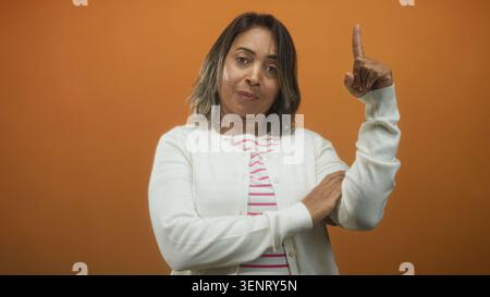 Die Frau zeigt den Zeigefinger nach oben, trägt im Studio eine weiße Strickjacke und ein gestreiftes Oberteil. Stockfoto