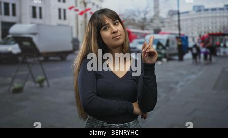 Die junge hispanische Frau zeigt den Zeigefinger nach oben mit gefaltetem Arm und sichtbarer Halskette auf der Straße in der Nähe von geparkten Lieferwagen und Fußgängern; ruhige Besinnung. Stockfoto