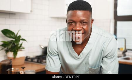 Lächelnder männlicher Healthcare-Mitarbeiter in Scrubs entspannend in heller Küche mit Kaffee und Pflanze Stockfoto