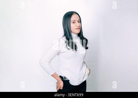 Junge südostasiatische brünette Frau vor isoliertem weißem Hintergrund, die mit verschränkten Armen steht, nach vorne schaut und subtil lächelt. Stockfoto