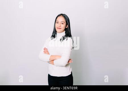 Junge südostasiatische brünette Frau vor isoliertem weißem Hintergrund, die mit verschränkten Armen steht, nach vorne schaut und subtil lächelt. Stockfoto