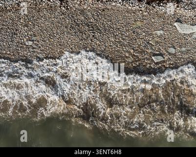 Drohnenblick von oben nach unten auf die Meereswellen, die an einem felsigen Kieselsteinstrand brechen. Stockfoto