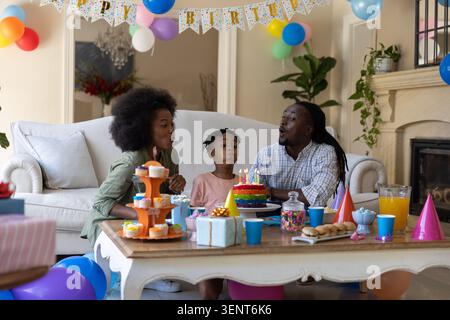 Afroamerikanische Familie, die Geburtstagskuchen ausbläst, während sie auf dem Sofa im Wohnzimmer sitzt Stockfoto