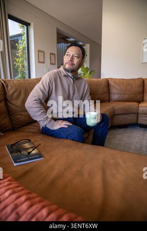 Reifer asiatischer Mann, der auf Ledersofa im Wohnzimmer sitzt und grünen Becher hält, Brille auf Buch Stockfoto