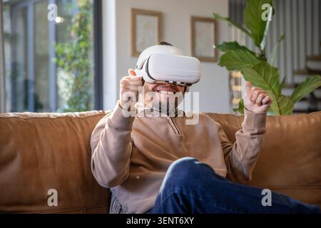 Asiatischer Mann lächelt und greift, während er ein weißes VR-Headset auf der braunen Couch im Wohnzimmer trägt Stockfoto