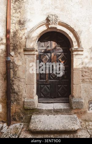 Nahaufnahme einer verwitterten, dunklen Holztür mit kunstvollen geometrischen Schnitzereien in einem rustikalen Kalksteinbogen in den historischen Gassen von Banon, Frankreich. T Stockfoto