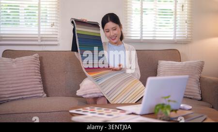 Junge asiatische Designerin präsentiert farbenfrohe Stoffmuster, während sie an der Möbelauswahl aus dem Home Office arbeitet Stockfoto