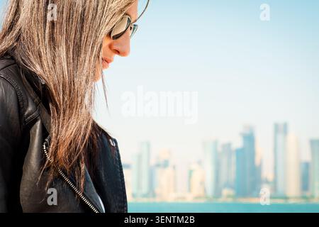 Nahaufnahme Profil junge Frau braune Haare, Sonnenbrille, schwarze Lederjacke. Unscharfe Skyline der Bucht, Hintergrund der Corniche am Wasser. Weiches Sonnenlicht, im Freien Stockfoto