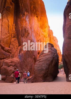 Touristen spazieren auf einem sandigen Boden zwischen massiven Felsbrocken in einem versteckten Canyon im Arches National Park, Moab, Utah Stockfoto