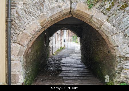 Blick durch einen schmalen, dunklen Steinbogentunnel mit Mauern, der eine leicht bergauf geschwungene Dorfstraße mit alten Steinbauten freigibt Stockfoto