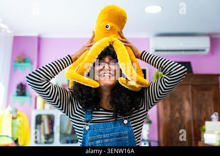 Junge hispanische Frau mit lockigen Haaren und Brille, die hell lächelt, verspielt einen gelben Kraken Plüsch auf dem Kopf hält und einen lässigen Moment genießt Stockfoto