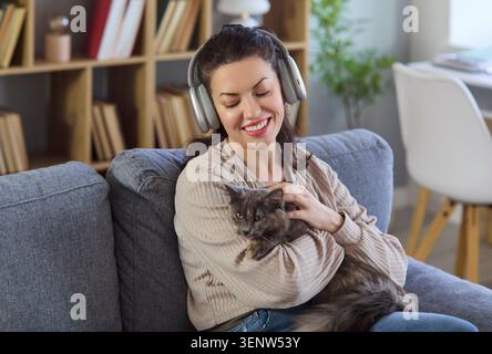 Lächelnde Frau in Kopfhörern kuschelt Katze auf Sofa zu Hause, hört Musik Stockfoto