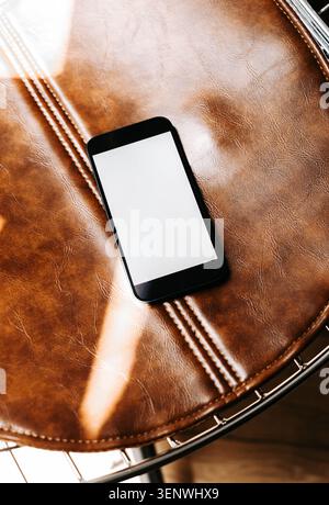 Smartphone mit leerem Bildschirm auf braunem Lederstuhl, moderner Technologie mit warmer Textur, glänzender Oberfläche und Kopierraum. Draufsicht Stockfoto