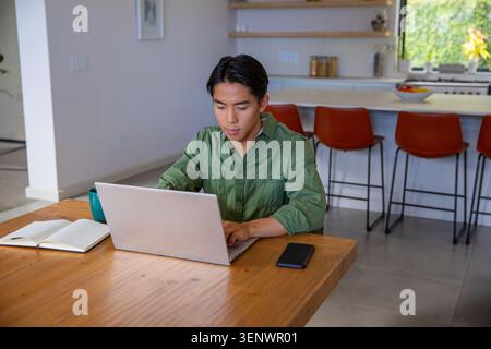 Erwachsener asiatischer Mann sitzt am Tisch in der modernen Küche, trägt ein grünes Hemd und tippt auf silbernem Laptop Stockfoto