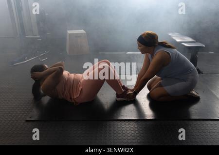 Zwei Erwachsene afroamerikanische Frauen trainieren auf schwarzer Matte, eine macht Sit-ups, Coach hält Knöchel Stockfoto