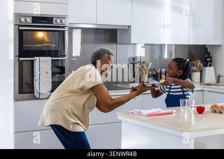 Eine ältere Frau, die als Jugendmädchen in Schürze auf der Kücheninsel ein Backblech hält Stockfoto