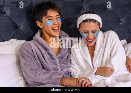 Verschiedene Paare lachen und Hände auf dem Bett halten, weiche Bademäntel und blaue Hydrogelflecken tragen Stockfoto