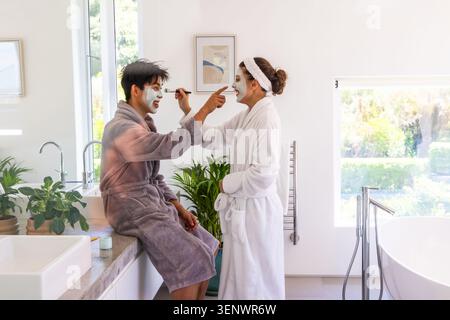 Diverse Paare tragen Bademäntel und Gesichtsmasken, Frauen, die Hautpflege mit Bürste auf der Arbeitsplatte anwenden Stockfoto