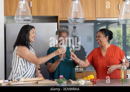 Verschiedene ältere Freundinnen stehen auf der Kücheninsel und knallen Weingläser von Baguette Stockfoto
