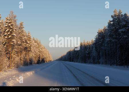 Die Straße schlängelt sich durch einen verschneiten Wald, während die Sonne untergeht und lange Schatten über den Boden wirft. Sein warmes, oranges Licht erfüllt die Szene mit einem Gefühl von... Stockfoto