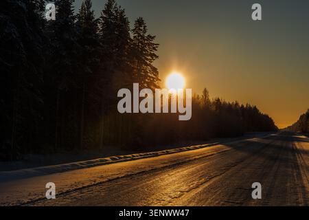 Die Straße schlängelt sich durch einen verschneiten Wald, während die Sonne untergeht und lange Schatten über den Boden wirft. Sein warmes, oranges Licht erfüllt die Szene mit einem Gefühl von... Stockfoto