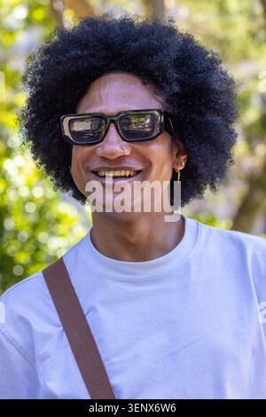 Erwachsener afrikanischer Mann lächelnd, trägt schwarze Töne, weißes T-Stück, Taschenriemen, Nasenbügel, Ohrring im Park Stockfoto