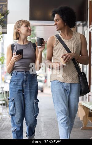 Diverse Paare, die auf dem Bürgersteig der Stadt laufen und Kaffeetassen, Smartphone, Rucksack, Umhängetasche halten Stockfoto