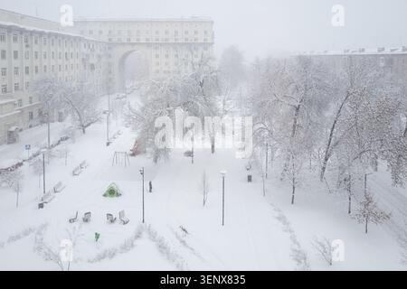 Ein ruhiger Stadtpark, der während eines Schneesturms von starkem Schneefall bedeckt ist, mit schneebedeckten Bäumen, einem Spielplatz und einem großen Apartmentgebäude. Stockfoto