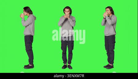 Person trinkt Kaffee vor der Kamera, hält eine Tasse heißes Koffeingetränk über einer Ganzkörper-Green-Screen-Vorlage, während der junge Mann Erfrischung genießt und sich im Studio positiv fühlt. Stockfoto