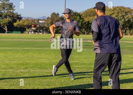 Männliche Teamkollegen werfen roten Cricket Ball, während Teenager-Teamkollegen in Hemden auf Gras bereit stehen Stockfoto