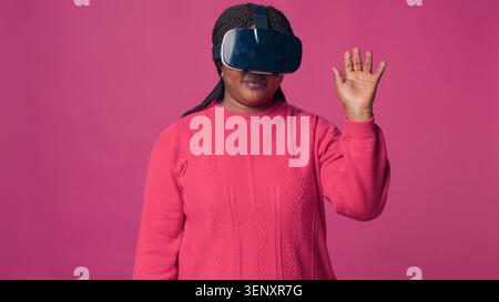 Innovative junge Frau genießt futuristische Simulation mit interaktivem Gerät, während sie im Studio ein VR-Headset trägt, und trendige Fashionista mit modernem VR-Headset. Stockfoto