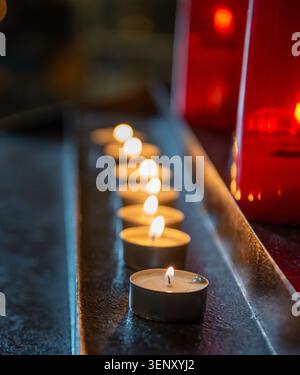 Beleuchtete Kerzen, in parallelen Reihen entlang einer dunklen Metall- oder Holzoberfläche angeordnet, typisch für Kirchenkerzenhalter. Stockfoto