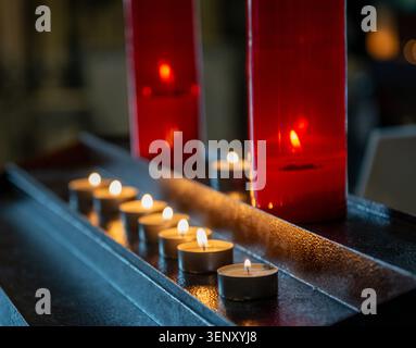 Beleuchtete Kerzen, in parallelen Reihen entlang einer dunklen Metall- oder Holzoberfläche angeordnet, typisch für Kirchenkerzenhalter. Stockfoto