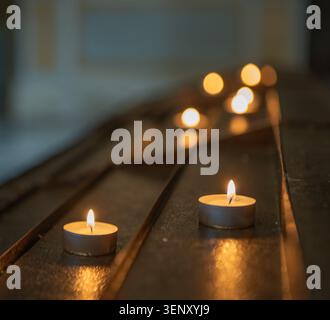 Beleuchtete Kerzen, in parallelen Reihen entlang einer dunklen Metall- oder Holzoberfläche angeordnet, typisch für Kirchenkerzenhalter. Stockfoto
