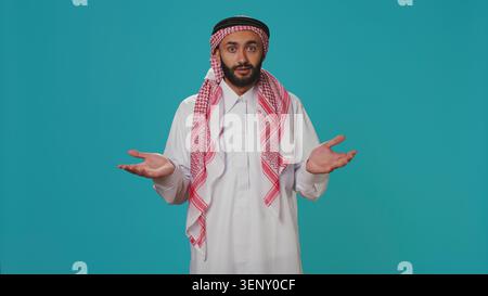 Islamischer Erwachsener, der sich im Studio unsicher und ahnungslos benimmt, das Zeichen „Ich weiß nicht“ zeigt und über blauem Hintergrund verwirrt ist, sich ein junger Mensch in arabischer Kleidung unsicher fühlt und zuckt. Stockfoto