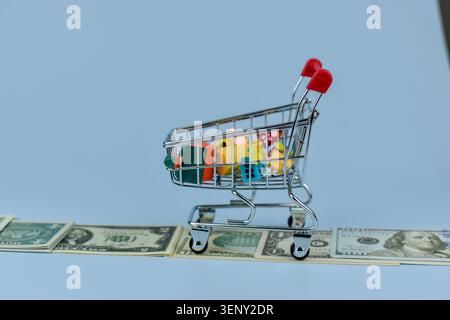 Der Mini-Einkaufswagen bietet Platz für bunte Spielzeuge. Dollars liegen unter dem Wagen auf glatter Oberfläche. Der hellrote Griff steht im Kontrast zum kühlen blauen Hintergrund. Symbolisiert Stockfoto
