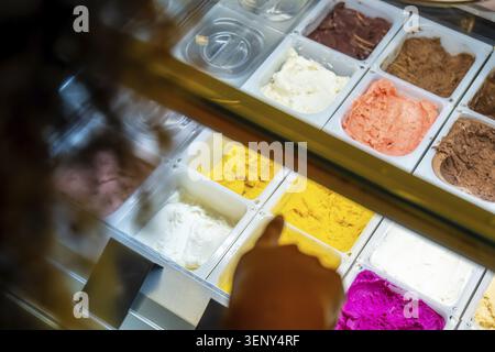 Kundenhand zeigt auf bunte Eis- und Sorbetaromen in einer gekühlten Vitrine, die Vielfalt und köstliche Dessert-Option hervorhebt Stockfoto