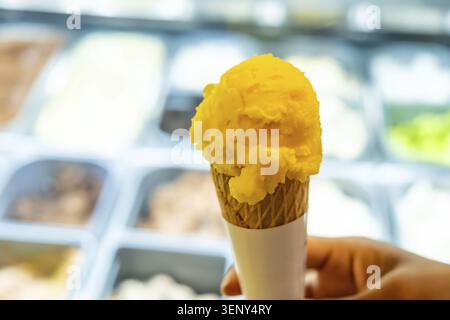 Halten Sie einen einzelnen Messlöffel lebhaftes gelbes Mangosorbet in einem Waffelkegel und bieten Sie eine erfrischende und köstliche Dessertoption aus einer Vitrine Stockfoto