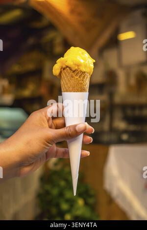 Die Hand hält einen einzelnen gelben Mangoeislöffel in einem Waffelkegel mit einem weißen Papierbeutel und genießt so einen süßen, cremigen Sommergenuss Stockfoto