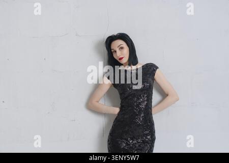 Stilvolle junge Frau mit schwarzen Haaren und roten Lippen trägt ein glitzerndes Cocktailkleid. Sie posiert selbstbewusst gegen eine minimalistische graue Betonwand. Hoch Stockfoto