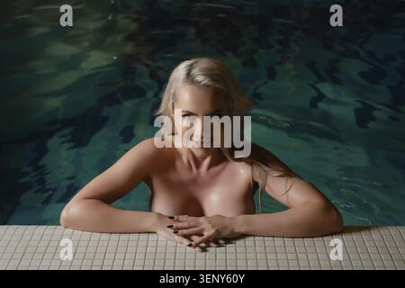 Eine Frau mit langen, blonden Haaren wird in einem Spa entspannt und sich sanft auf eine gekachelte Kante lehnt. Die ruhige Atmosphäre wird durch einen ruhigen Pool, Einladungen, verstärkt Stockfoto