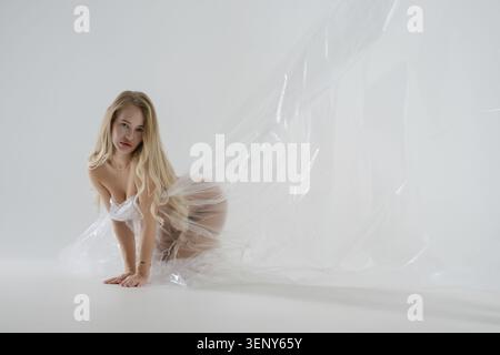 Eine schöne blonde Frau posiert in einem Schönheitsstudio und kniet mit fließenden transparenten Stoffen, die um sie herum hängen Stockfoto