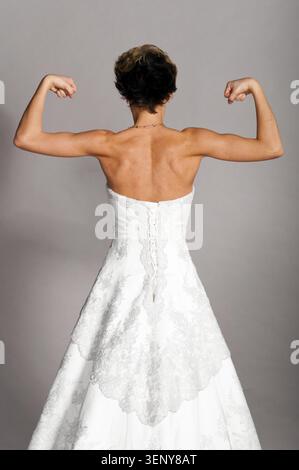 Die schöne Braut steht und zeigt ihre starken Hände im Hochzeitskleid auf grauem Hintergrund, Blick von hinten Stockfoto