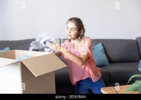 Frau kniet neben einem offenen Karton im Wohnzimmer, inspiziert Luftpolsterfolie, trägt rosa Oberteil Stockfoto
