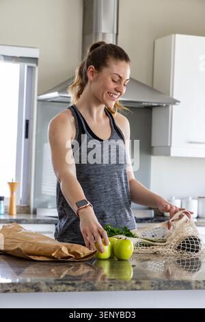 Nach dem Einkaufen, Frau im grauen Tank in der Küche, die grüne Äpfel in einen Netzbeutel packt und das Essen vorbereitet Stockfoto
