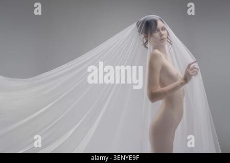 Eine selbstbewusste Frau posiert anmutig in zartem, transparentem Stoff und unterstreicht ihre Schönheit in einem eleganten Innenbereich Stockfoto